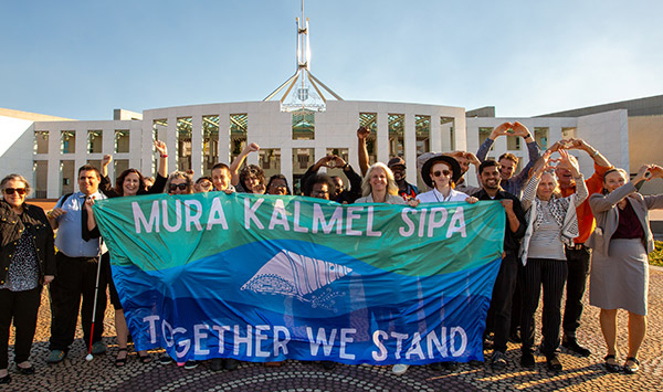 A large group pf people standing in front of Australia's Parliament House holding a banner that says Mura Kalmel Sipa, Together we stand.